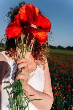Selfie of a curly red-haired woman covering her face with a bunch of red field poppies among a poppy field.