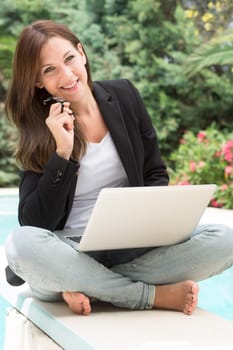 A young woman in blazer and glasses sits outside on the laptop