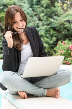 A young woman in blazer and glasses sits outside on the laptop