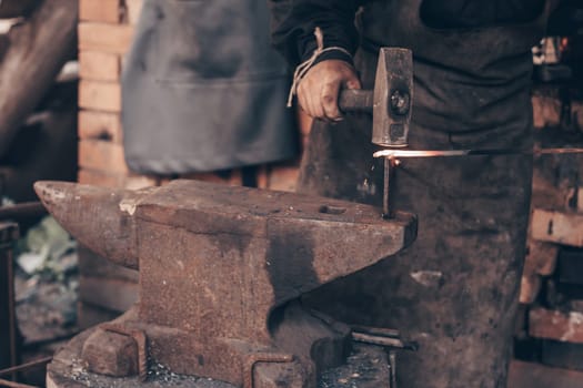 Blacksmith process glowing metal with hammer on anvil in forge. Dirty hands of farrier strike iron in workshop. Metalworking, manufacturing equipment and tools. Traditional technic of making hardware.
