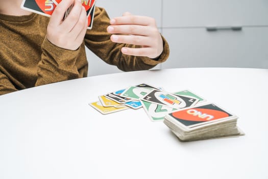Belarus, Minsk, 08.01.2023. Boy play UNO sitting at white table. Hands of child put paper colorful cards on top of each other. Interesting card game. Home leisure, playtime. Board game for kids.
