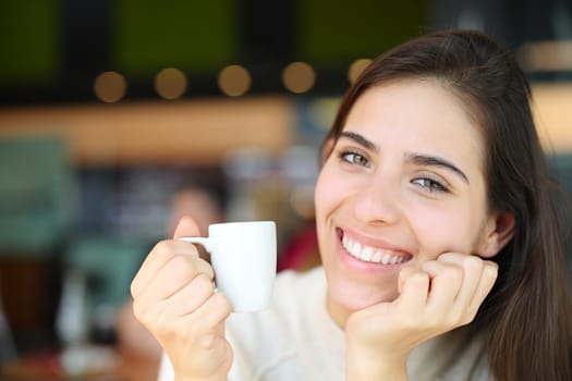 Happy woman holding cofee looks at you sitting in a bar