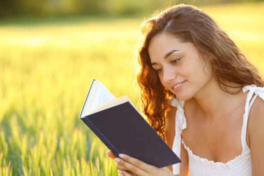 Happy woman sitting in a field reading a paper book at sunset