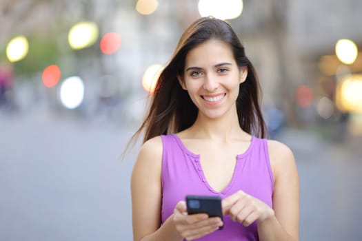 Front view portrait of a happy woman holding smart phone in the street looking at you