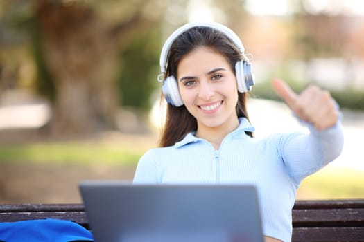 Happy student with laptop and headphone with thumbs up on a bench in a park
