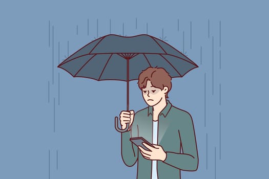 Sad man with umbrella stands in rain and reads SMS in mobile phone from girlfriend who refused to come on date. Concept of autumn depression due to bad weather and weather sensitivity