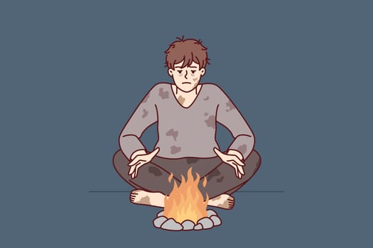 Homeless man in dirty clothes warms hands sitting by fire trying to survive due to lack of own home. Homeless guy needs help from social services and benefits or food stamps after losing job