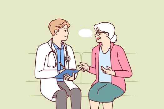 Elderly woman with practitioner doctor sit on couch complaining to hospital employee about health problems and tells symptoms of disease. Man doctor visits patient at home after pensioner call to 911
