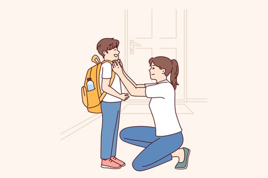Single mother prepares young son of student in elementary school as trip to lessons and helps to put on briefcase kneeling down. Caring mother dresses teen boy for first day of school