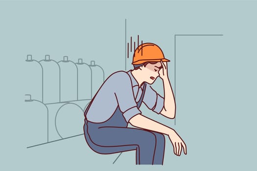 Tired man working in factory sits near production equipment holding head because of nervous job. Nervous factory worker in safety helmet needs psychological support due to poor working conditions