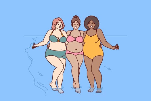 Cheerful plus size women dressed in swimsuit stand hugging on sunny beach near sea water and look at camera smiling. Diverse plus size girls for concept of no prejudice and body positivity