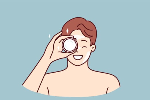 Man demonstrates jar of cream bought in cosmetic store to moisturize his face or get rid of wrinkles. Young guy with bare shoulders recommends purchasing nourishing balm or cream for skin care