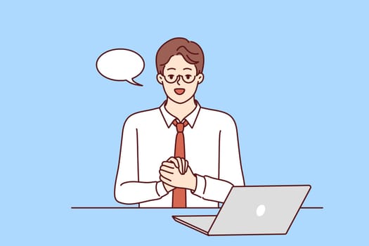Confident businessman is sitting at table with laptop and looking at camera recording business training. Guy in business clothes with dialogue cloud works as teacher online training courses