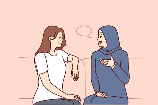 Girls from different ethnicities are talking sitting on couch demonstrating tolerance and lack of cultural prejudice. Woman in muslim clothes is talking to girlfriend of european appearance