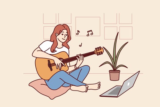 Woman plays guitar sitting in front of laptop and gets musical skills from watching video lesson on internet. Girl enjoys musical hobby and performance of popular compositions on guitar