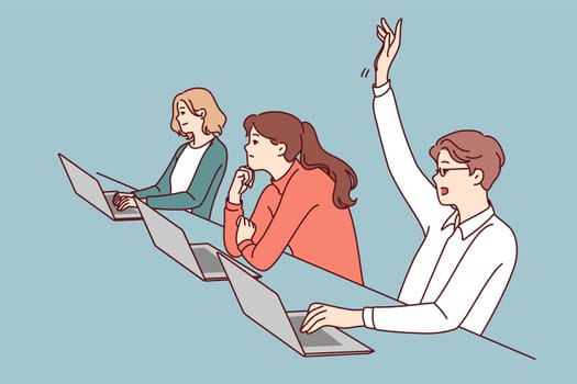Man student sitting at desk with laptop raises hand to attract attention of teacher and ask question on topic of lecture. People listening to lecture in college or university auditorium