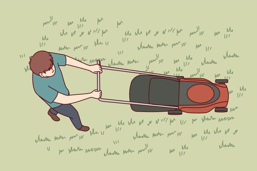 Man uses lawn mower to cut grass and wild weeds on golf course in park or in own backyard. Top view of young guy dressed in casual clothes with lawn mower working as gardener or janitor.