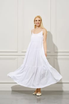 A beautiful happy woman in a waving white sundress stands elegantly indoors