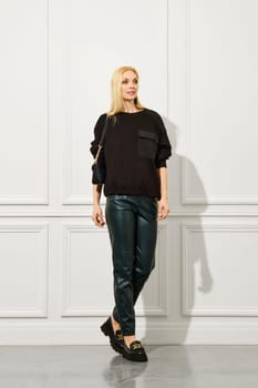 Cheerful woman stands in leather trousers and pullover in bright room. From head to toe she is dressed casually but fashionably with their footwear completing the look.
