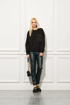 Smiling, a stylish woman poses indoors wearing trendy leather boots, slim trousers and a jumper with her blonde hair in urban fashion.