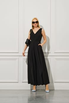 Stylish blonde businesswoman in black dress and sunglasses holds handbag on her bare shoulder