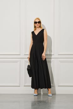 Confident businesswoman in long black dress and sunglasses stands in a bright room holding little handbag