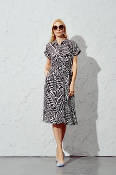 A stylish woman walking forward in a patterned dress and sunglasses under harsh sunlight, looking directly at the camera