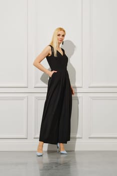 A blond businesswoman stands in a bright room, dressed fashionably in long black dress and wearing stilettos. She holds one hand in a pocket