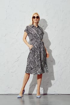 A smiling blonde woman in a patterned dress and sunglasses stands confidently harsh sunlight, looking directly at the camera