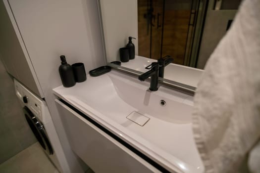 Modern bathroom sink with black matte faucet and minimalist accessories. White countertop and a towel hang nearby.