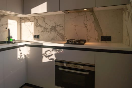 Modern kitchen with marble backsplash, built-in oven, gas cooktop, and sleek gray cabinetry. Sunlight streams in.