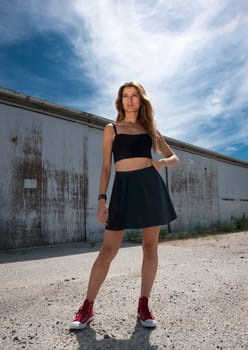 Full length low angle front view of pretty fashion model wearing short black skirt, standing outdoors in grunge setting