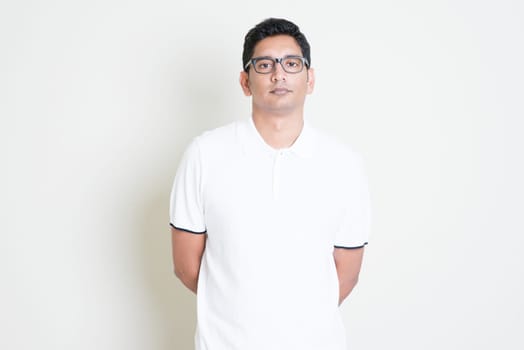 Portrait of bored Indian guy with serious face expression. Asian man standing on plain background with shadow and copy space. Handsome male model.