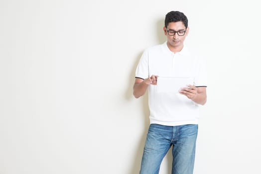 Portrait of handsome Indian guy using tablet pc, standing on plain background with shadow, copy space at side.