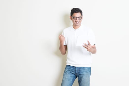 Portrait of excited Indian guy using tablet pc and celebrate his success . Asian man standing on plain background with shadow and copy space. Handsome male model.