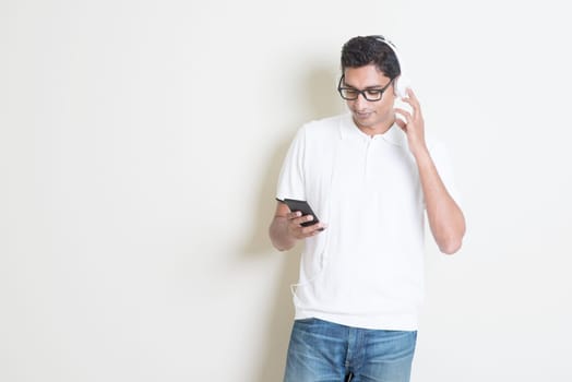 Smartphone social media concept. Indian guy using mobile phone. Asian man relaxed and listening music with headset, standing on plain background. Handsome male model.
