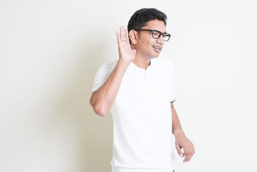 Portrait of Indian guy listening, hand cupped beside ears. Asian man standing on plain background with shadow and copy space. Handsome male model.
