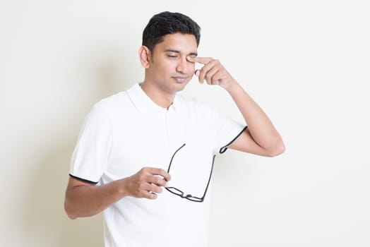 Portrait of Indian guy take off eyeglasses and rubbing his tired eyes. Asian man standing on plain background with shadow and copy space. Handsome male model.
