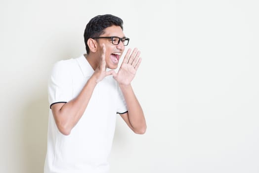 Portrait of Indian guy shouting loud, mouth open and hand cupped beside. Asian man standing on plain background with shadow and copy space. Handsome male model.