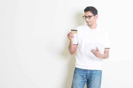 Portrait of Indian guy shopping online using tablet pc, doing transaction by checking credit card. Asian man standing on plain background with shadow and copy space. Handsome male model.