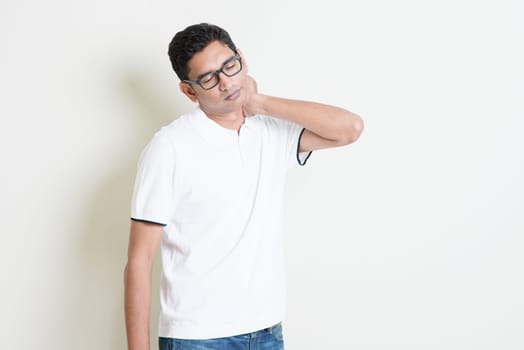 Portrait of Indian guy neck pain, massaging with tired face expression. Asian man standing on plain background with shadow and copy space. Handsome male model.