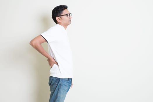 Portrait of Indian guy backache, holding spine with hand. Asian man standing on plain background with shadow and copy space. Handsome male model.