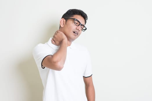 Portrait of tired Indian guy massaging his neck with painful face expression. Asian man standing on plain background with shadow and copy space. Handsome male model.