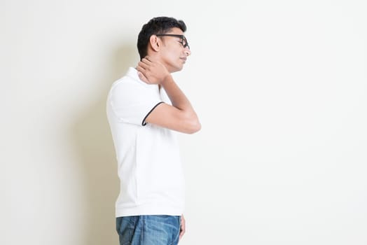 Portrait of Indian guy neck pain, massaging with hand. Asian man standing on plain background with shadow and copy space. Handsome male model.