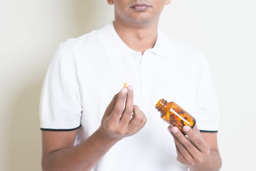 Sick Indian guy taking medicine capsule. Asian man standing on plain background with shadow and copy space.