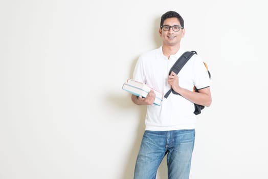 Portrait of adult Indian university student with books and bag. Asian man standing on plain background with shadow and copy space. Handsome mixed race male model.