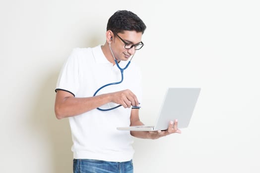 Spyware or malware concept. Indian computer engineer diagnose virus infected pc with stethoscope. Asian man standing on plain background with shadow and copy space.