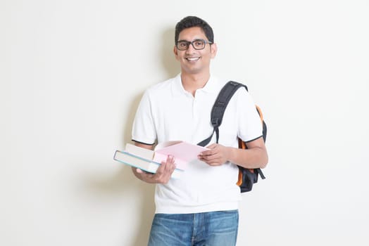 Portrait of adult Indian college student with bag and books. Asian man standing on plain background with shadow and copy space. Handsome mixed race male model.