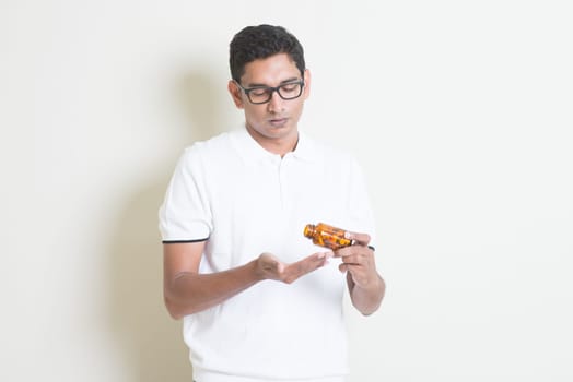 Sick Indian guy taking tablet medicine. Asian man standing on plain background with shadow and copy space.