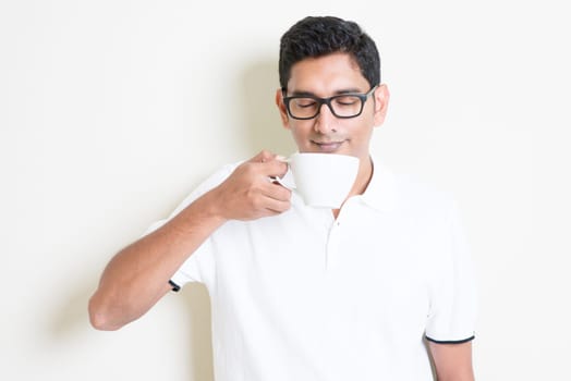 Indian guy holding a cup, drinking and enjoying the aroma coffee. Asian man standing on plain background with shadow and copy space. Handsome male model.